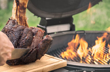 Beefhammer van de schenkel van het Grainfed Vaars rundvlees wordt aangesneden naast een brandende barbecue.