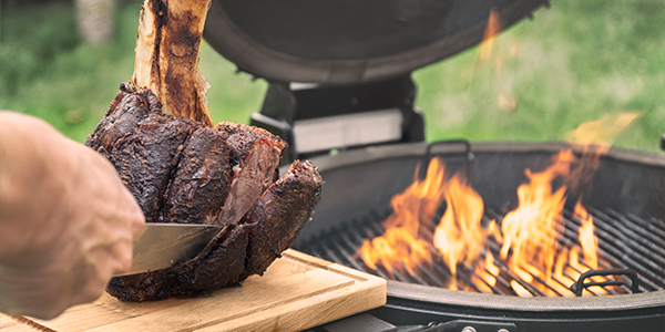 Beefhammer van de schenkel van het Grainfed Vaars rundvlees wordt aangesneden naast een brandende barbecue.